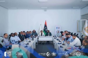 FB_IMG_1781127235761 - كلية الصيدلة - جامعة سبها Group of officials seated around a U-shaped conference table in a formal meeting room, with a Libyan flag at the center and banners in the background.