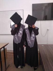 Two graduates in black gowns and gray sashes stand indoors, faces obscured by oversized square mortarboards.