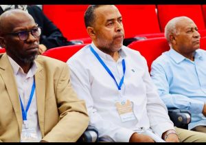 Three men sit in a row at a conference, each wearing a blue lanyard with an ID badge; red seats surround them.