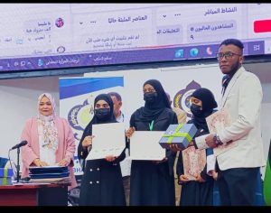 Group of graduates receiving certificates at an awards ceremony; four women in hijabs and masks stand with a man in a light suit holding gifts on stage.