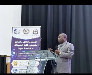 Man in a gray suit giving a speech at a glass podium on a stage, with a purple banner and logos to the left written in Arabic behind him.