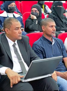 Two men sit in a conference hall with red seats; one in a suit using a laptop, the other in a polo shirt, while background attendees wear masks.