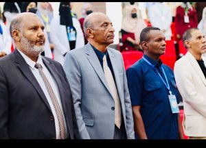 Four men in formal attire stand in a row at an event, with attendees and red seats blurred in the background.