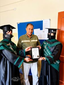 Two graduates in cap and gown present a framed certificate to a man in a green striped shirt during a ceremony in a classroom setting.