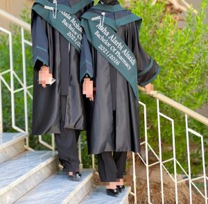Three graduates in black gowns with green sashes and caps pose on outdoor stairs, faces blurred for privacy.—graduation scene