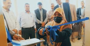 Woman in a hijab and mask cuts a blue ribbon with scissors at a ribbon‑cutting ceremony, surrounded by onlookers in business attire.