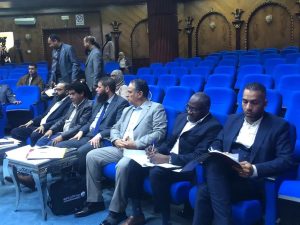 Group of men in business suits seated in blue theater seats, some taking notes, in an ornate auditorium with wood-paneled walls.