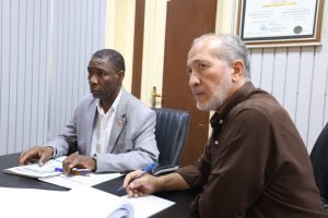 Two men seated at a desk reviewing documents during a meeting, one in a light gray suit and the other in brown shirt in an office setting with papers and pens on the table.