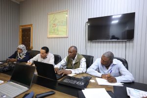 Four people sit at a conference table in a gray-paneled room, each with a laptop or notebook, focused on work in a meeting setting.
