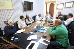 A group of diverse professionals seated around a long conference table in a meeting room, papers, notebooks, cups, and smartphones spread across the table as they discuss.