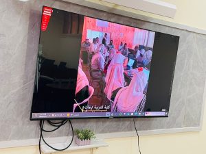 TV screen mounted on a marble wall showing a group of people in a meeting, many wearing hijabs, around a conference table; Arabic caption at bottom.