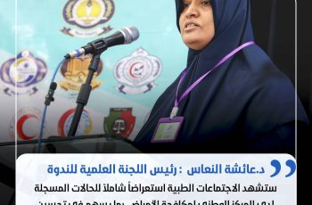 Woman in a navy hijab speaking at a podium with a microphone, wearing a purple ID badge on a lanyard.