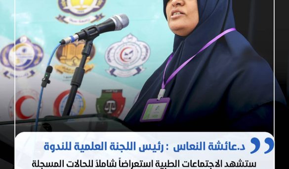 Woman in a navy hijab speaking at a podium with a microphone, wearing a purple ID badge on a lanyard.