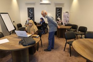 Man leans over a hijab-clad woman working on a laptop at a round table in a small training room.