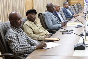Six men seated at a conference table with microphones, attentively listening during a meeting in a boardroom.