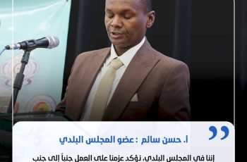 Man in a brown suit speaking at a podium with a microphone during a formal event, backdrop banner visible behind him, and a blue quotation overlay in Arabic text.