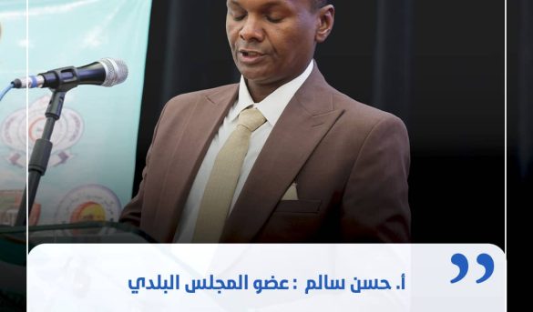 Man in a brown suit speaking at a podium with a microphone during a formal event, backdrop banner visible behind him, and a blue quotation overlay in Arabic text.