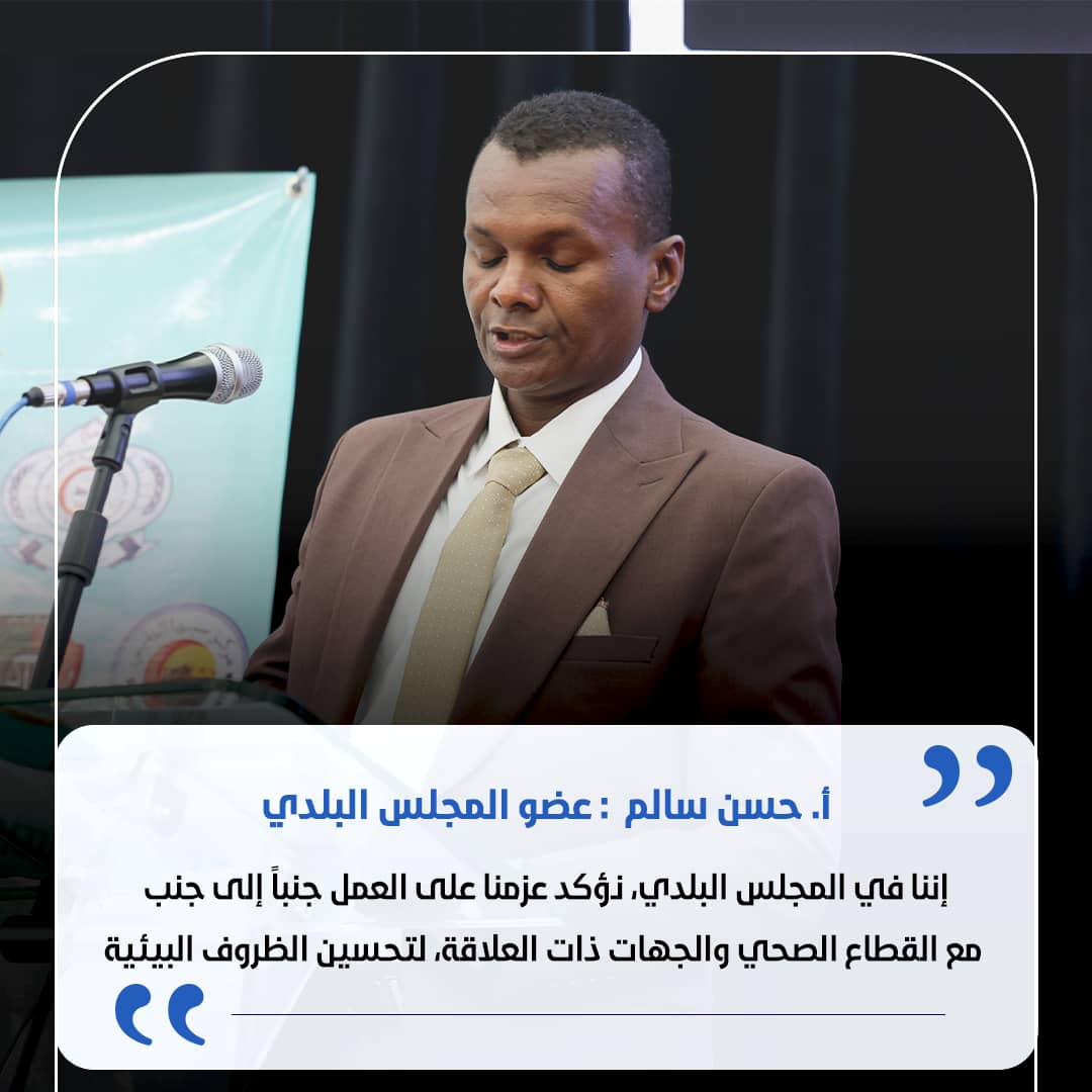 Man in a brown suit speaking at a podium with a microphone during a formal event, backdrop banner visible behind him, and a blue quotation overlay in Arabic text.