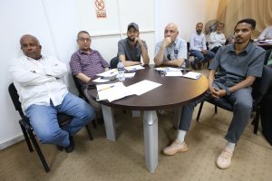 Group of men seated around a round table in a meeting room, papers and pens spread across the table as they listen.