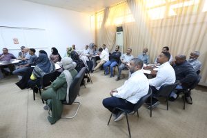 A diverse group of adults seated around tables in a meeting room, listening and taking notes.