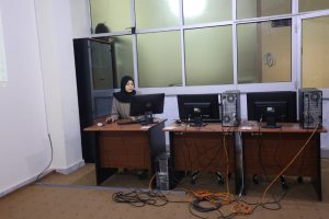 Woman in hijab sits at a desk with a monitor in a server-filled computer lab; tangled cables on the floor.