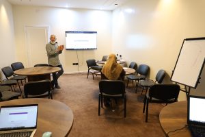 36019EE6-4E9F-4329-9E43-38AB95BF5491 - جامعة سبها | Sebha University Presenter at front of a small classroom, gesturing beside a mounted screen as a student in a hijab sits at a round table.