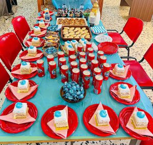 Long party table set with red plates and bright blue cupcakes, turquoise tablecloth, and center candy bowl; many red Fanta cans line the table edges.