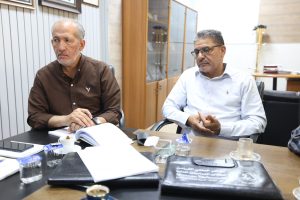Two middle-aged men sit at a conference table in an office, discussing with papers and beverages on the table in front of them.