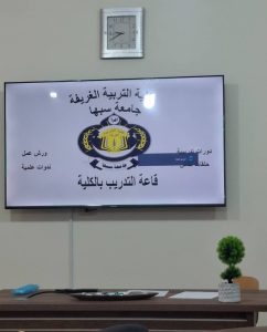 TV screen showing Arabic university emblem and text, in a classroom setting with a desk and a small potted plant nearby.