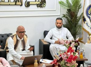 41a8d45f-585b-4f88-a3e1-10bd11e8f68c - جامعة سبها | Sebha University Two men in traditional white robes sit in a conference room with a laptop, papers, and a pink/red floral arrangement on the table.