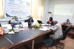 Panel-style meeting around a wooden conference table in an office; four people sit with name cards, a banner in Arabic on the wall, and a projector screen in the background.