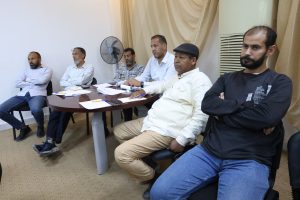 Six men sit at a round table in a meeting room, papers and pens spread across the surface.