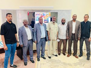 Eight men in business attire stand in a row in a conference room, with a large screen on the wall behind them.