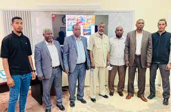 Eight men in business attire stand in a row in a conference room, with a large screen on the wall behind them.