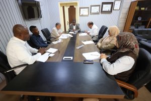 Seven professionals sit around a wood conference table reviewing papers together in a meeting room.