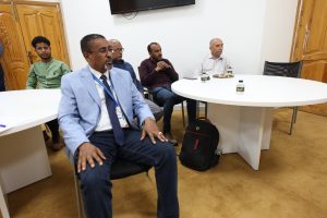 Group of men seated around a white round conference table in a meeting room, some with drinks, looking attentive to something off-camera.