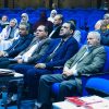 Audience seated in a blue theater, watching a formal presentation in suits and hijabs.