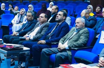 Audience seated in a blue theater, watching a formal presentation in suits and hijabs.