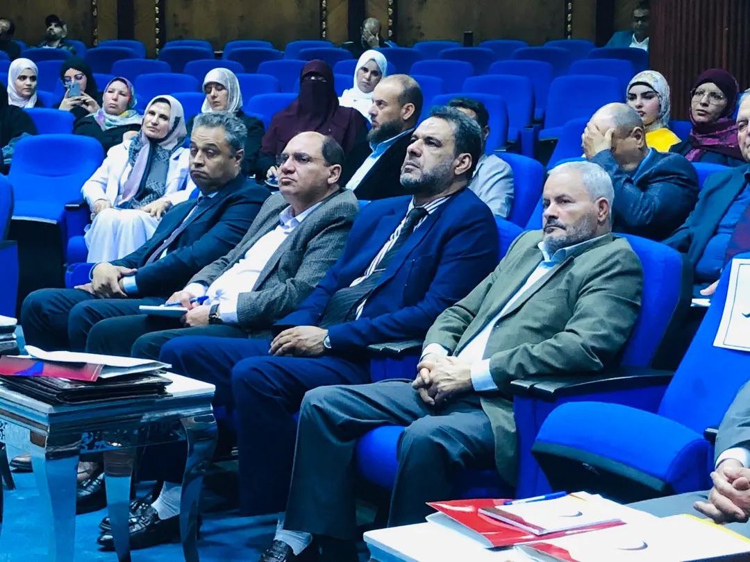 Audience seated in a blue theater, watching a formal presentation in suits and hijabs.