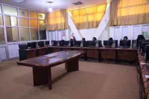 A computer lab with many stations along two long desks, people wearing hijabs using desktop PCs, central wooden table in the foreground, yellow curtains in the background.