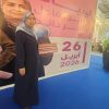 Woman in a dark blue abaya and hijab stands on a blue floor, holding a stack of books next to a large pink event banner with the date 2026.