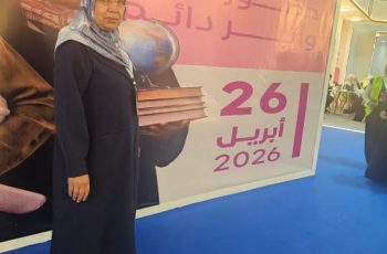 Woman in a dark blue abaya and hijab stands on a blue floor, holding a stack of books next to a large pink event banner with the date 2026.
