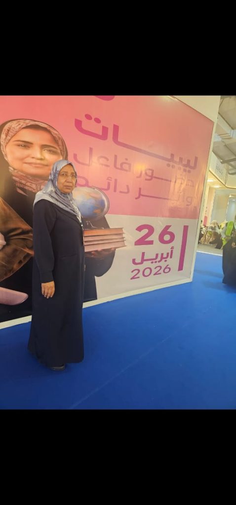 Woman in a dark blue abaya and hijab stands on a blue floor, holding a stack of books next to a large pink event banner with the date 2026.