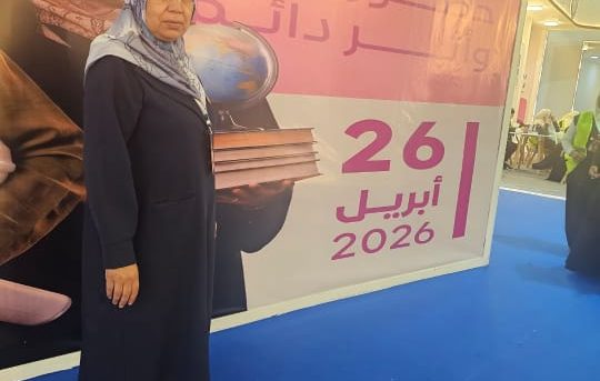 Woman in a dark blue abaya and hijab stands on a blue floor, holding a stack of books next to a large pink event banner with the date 2026.