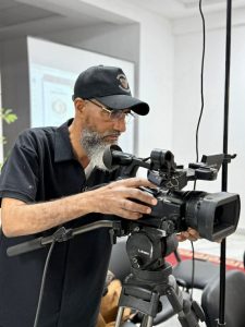 5f4baa08-f360-4933-9fb1-3b5702297804 - جامعة سبها | Sebha University Man with a beard operating a professional video camera mounted on a tripod indoors, wearing a black cap and polo shirt.