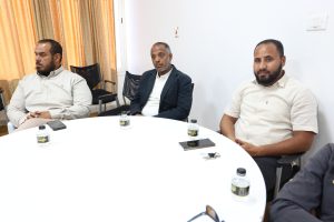Three men sit around a white round table in a meeting room, with water bottles and phones on the table and orange curtains in the background.