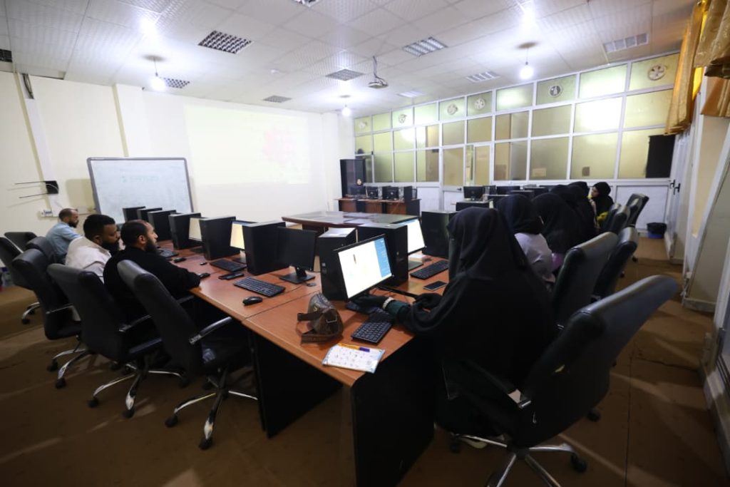 Group of students (many wearing hijabs) sit at a U-shaped computer lab with monitors and a projector in front of them and a whiteboard to the side, learning session ongoing in a classroom.