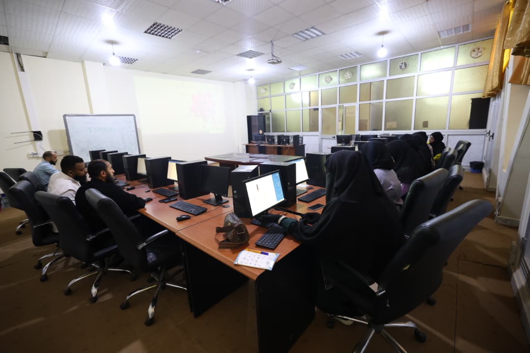 Group of students (many wearing hijabs) sit at a U-shaped computer lab with monitors and a projector in front of them and a whiteboard to the side, learning session ongoing in a classroom.