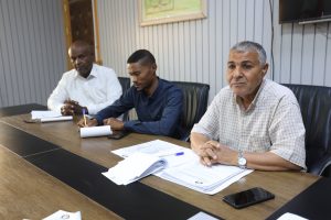 Three men sit at a conference table in a meeting room, reviewing documents and notes.