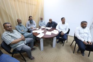 A group of seven men sit around a round table in a meeting room, papers and pens scattered on the table, beige curtains to the left and a plain white wall to the right.